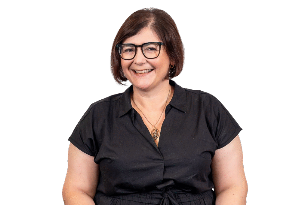 A woman with short brown hair and glasses smiles while wearing a black short-sleeve shirt and gold necklace, posed against a plain white background—perfect to represent music therapy courses or a music therapist Gold Coast professional.