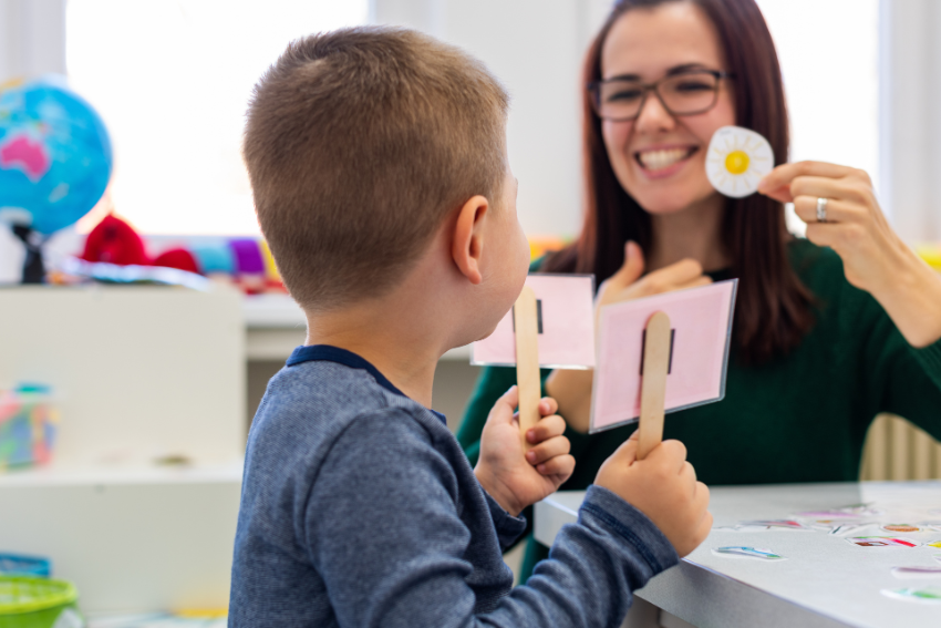 A young boy in a classroom holds up cards on sticks while a smiling adult woman shows him a card with a picture of a flower, engaging in an interactive speech therapy for kids session.