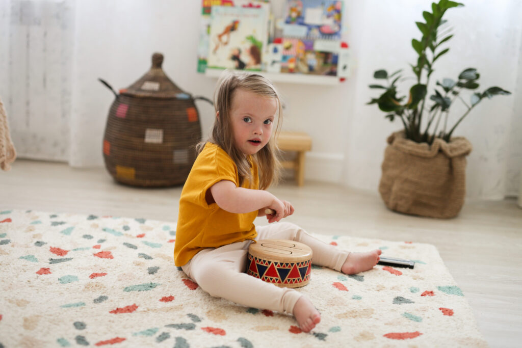 A young girl with Down syndrome sits on a patterned rug, playing a small drum—an activity often used in music therapy for kids. She wears a yellow shirt and light pants in a bright, cozy room filled with toys, plants, and shelves.