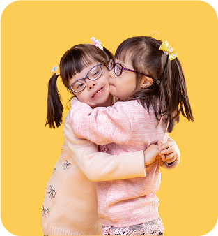 Two young girls with glasses and pigtails, wearing pastel sweaters, hug each other closely and smile in front of a solid yellow background—radiating the warmth of friendship found in music therapy gold coast sessions.