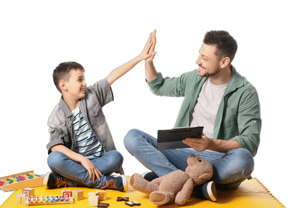 A man and a boy sitting on a yellow mat give each other a high-five. The man holds a tablet, and toys and a teddy bear are in front of them. Both are smiling, enjoying their time together—perhaps during speech therapy for kids.