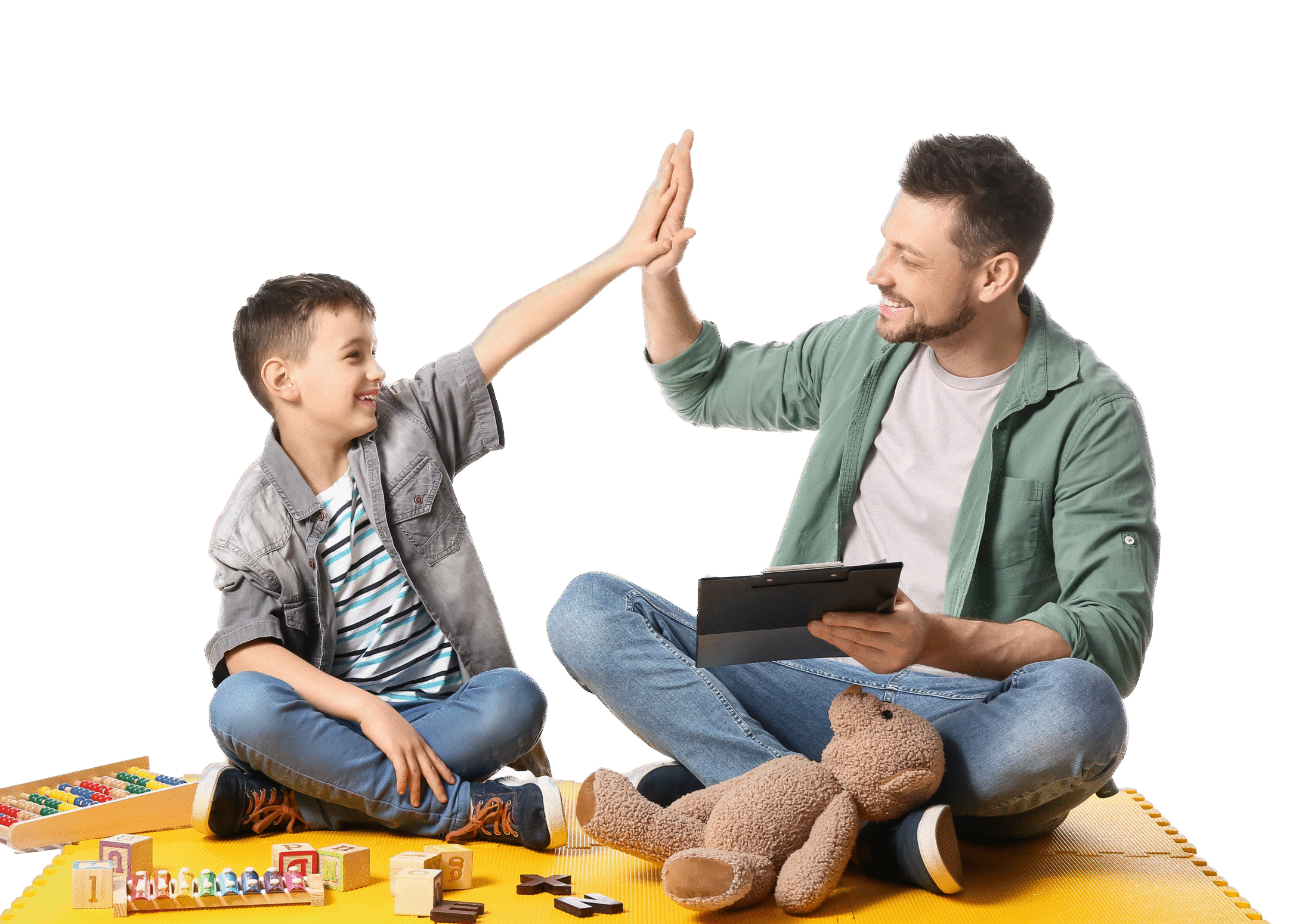 A man and a boy sitting on a yellow mat give each other a high-five. The man holds a tablet, and toys and a teddy bear are in front of them. Both are smiling, enjoying their time together—perhaps during speech therapy for kids.