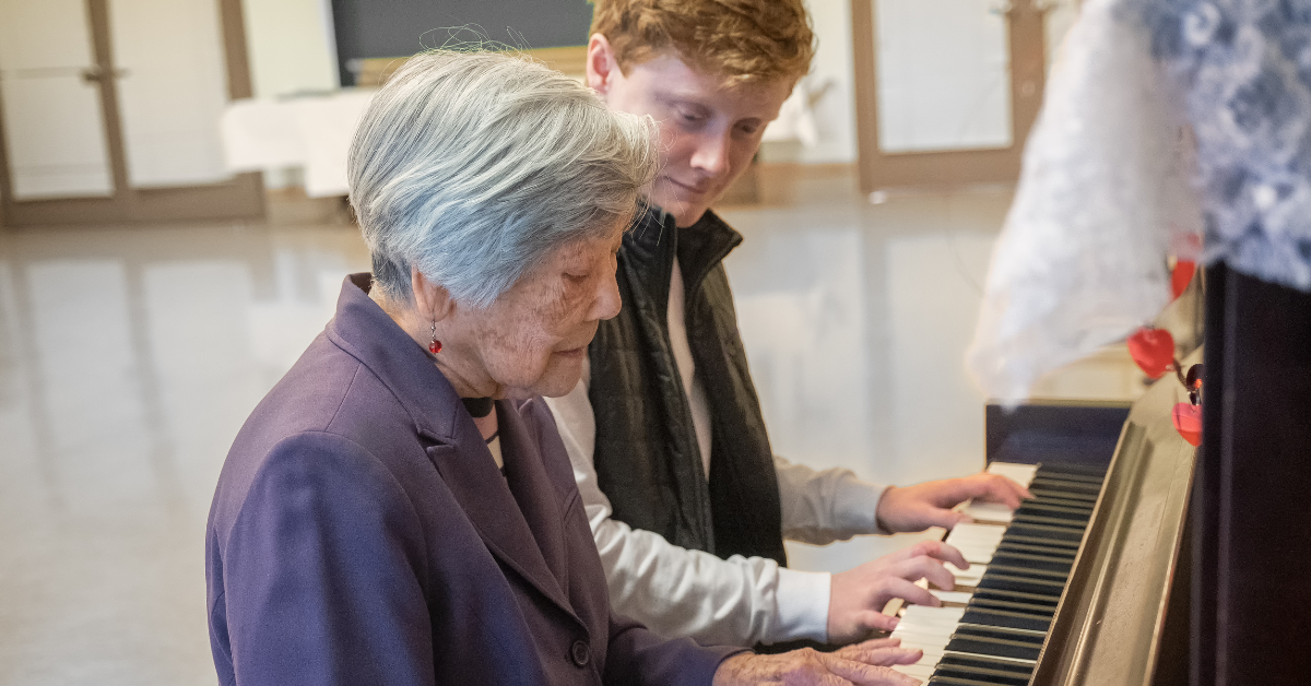 An elderly woman and a young man play piano together, sitting side by side. Sharing a musical moment, they focus on the keys—showcasing the power of music therapy gold coast to unite generations through creativity and connection.