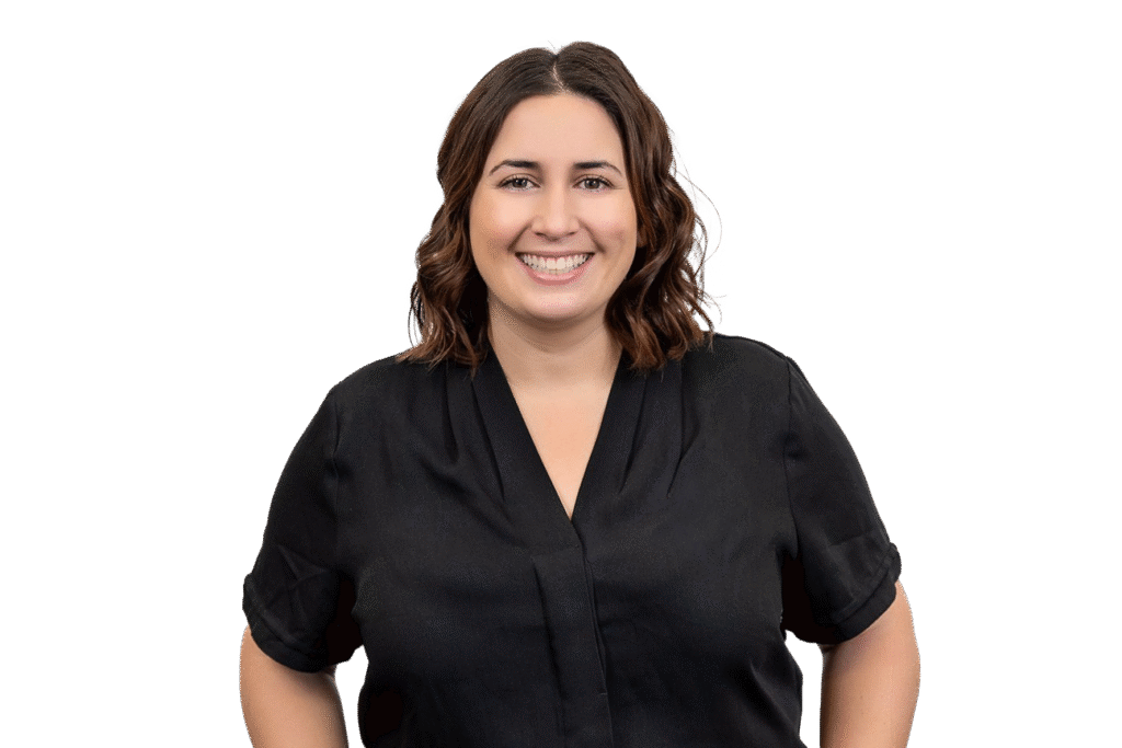 A woman with wavy brown hair, wearing a black short-sleeve blouse, smiles while standing against a plain white background—a welcoming presence for anyone interested in music therapy Gold Coast services.