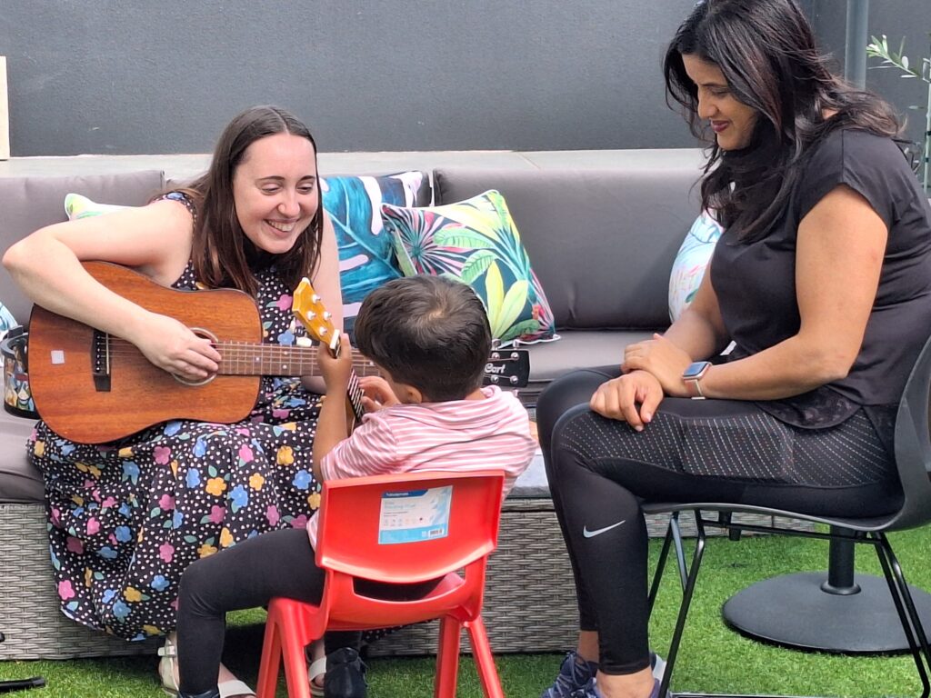 a woman playing guitar in a music therapy session with a young boy and his mother