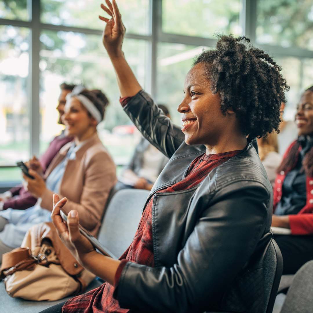 Woman sitting with her hand up to ask a question with other people seated in the background.