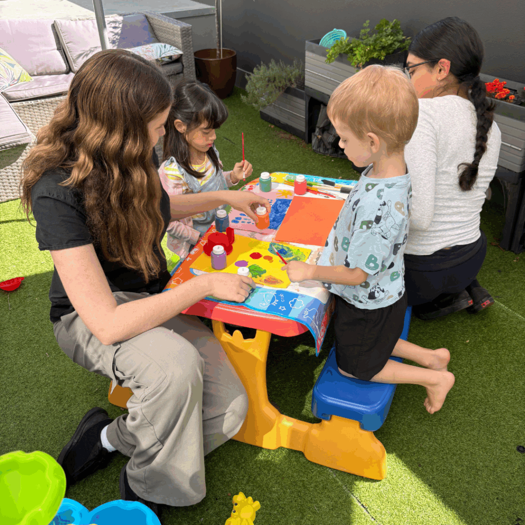 Two children sitting with two adult women at table outside, painting.
