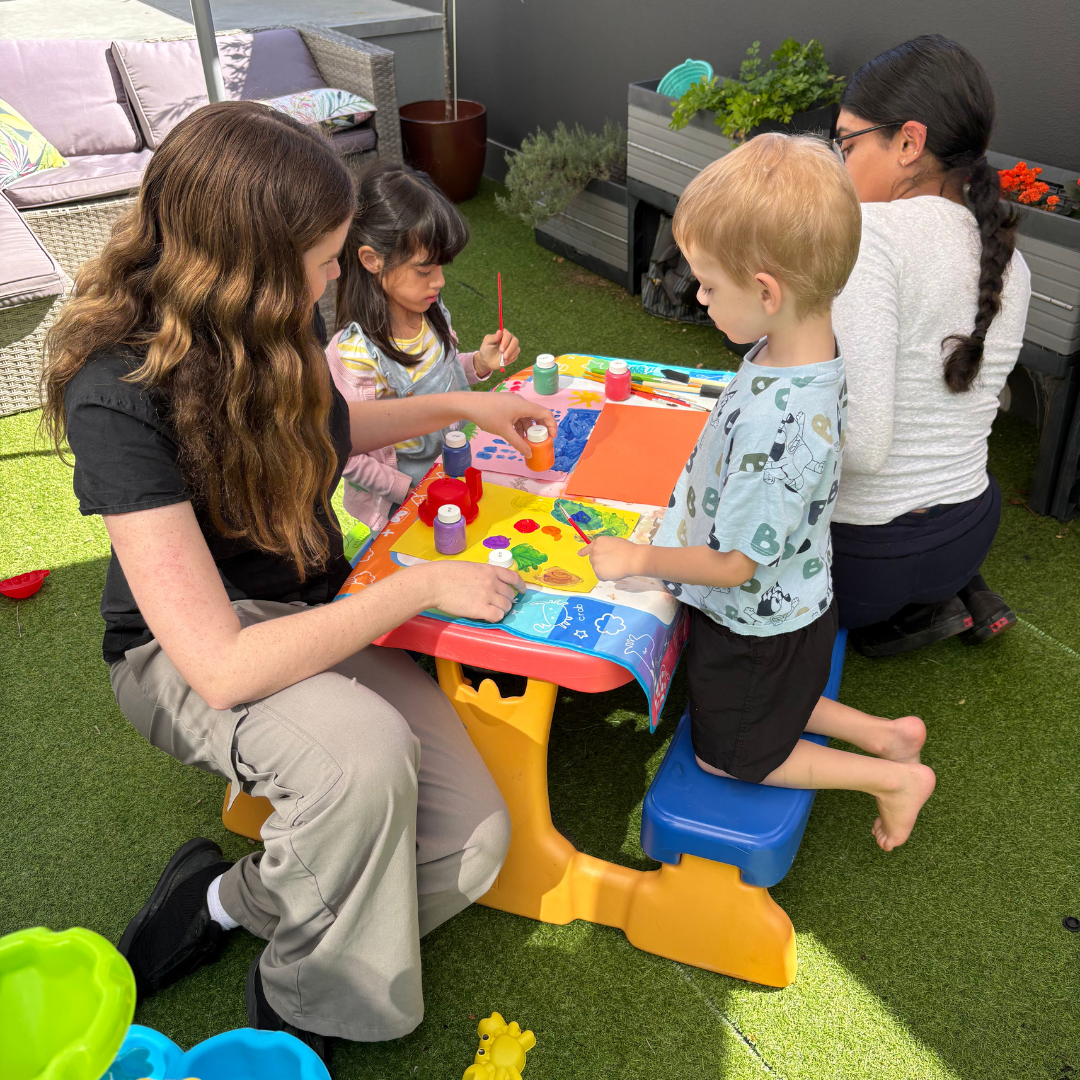 Two children sitting with two adult women at table outside, painting.