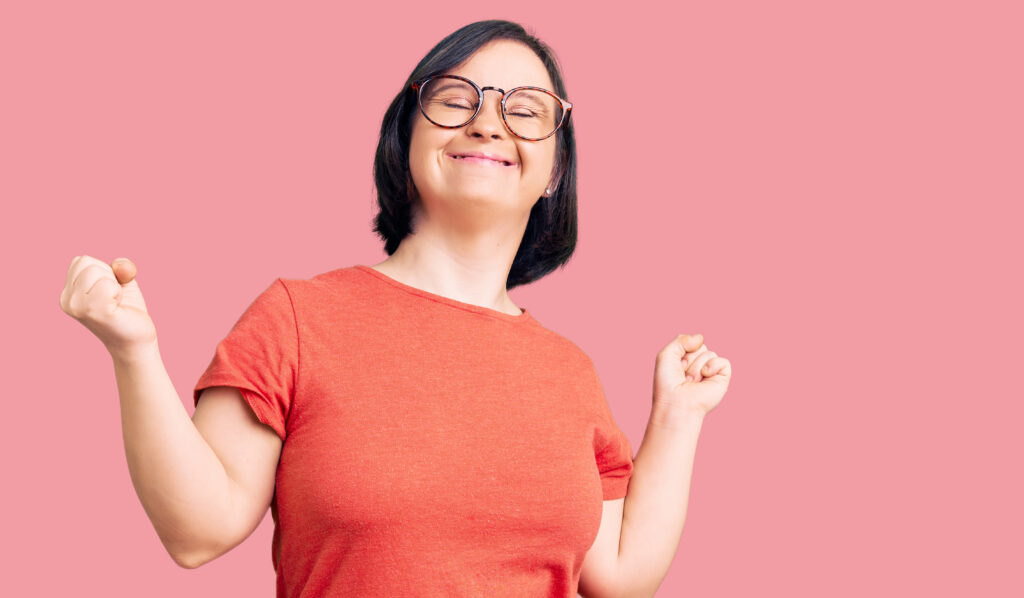 A woman with short dark hair and glasses smiles joyfully with eyes closed and arms raised, wearing an orange shirt, standing against a solid pink background—capturing the joy found through music therapy courses.