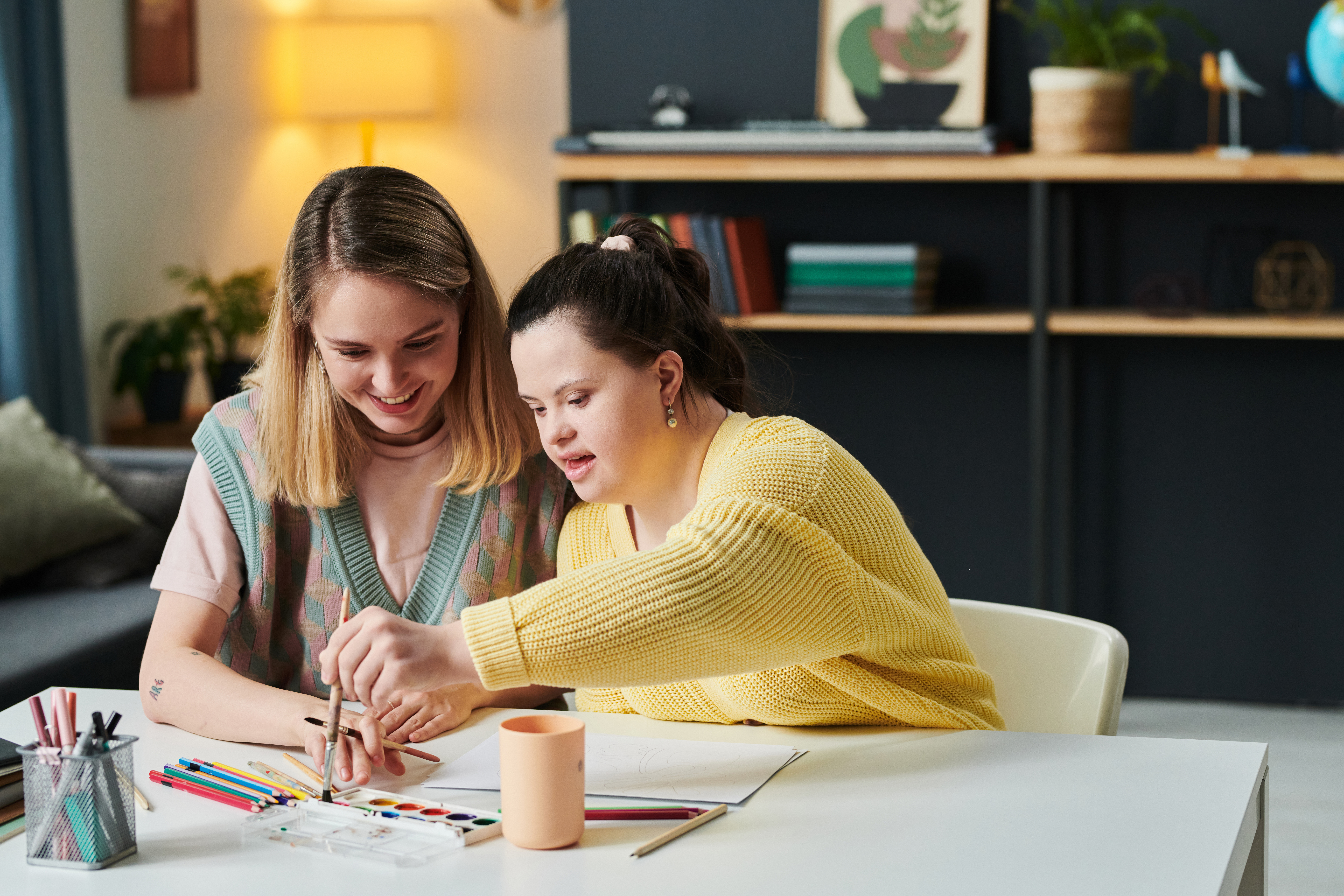 Two young women sit at a table, smiling and painting with watercolours as an OT Gold Coast therapist gently guides one woman’s hand. Coloured pencils and paper are spread out before them in a cosy, well-lit room.