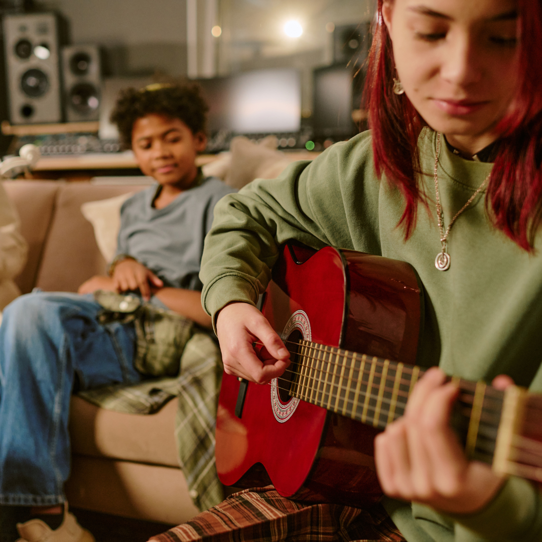A close-up photo of a teenage girl playing guitar with another girl in the background listening to her.