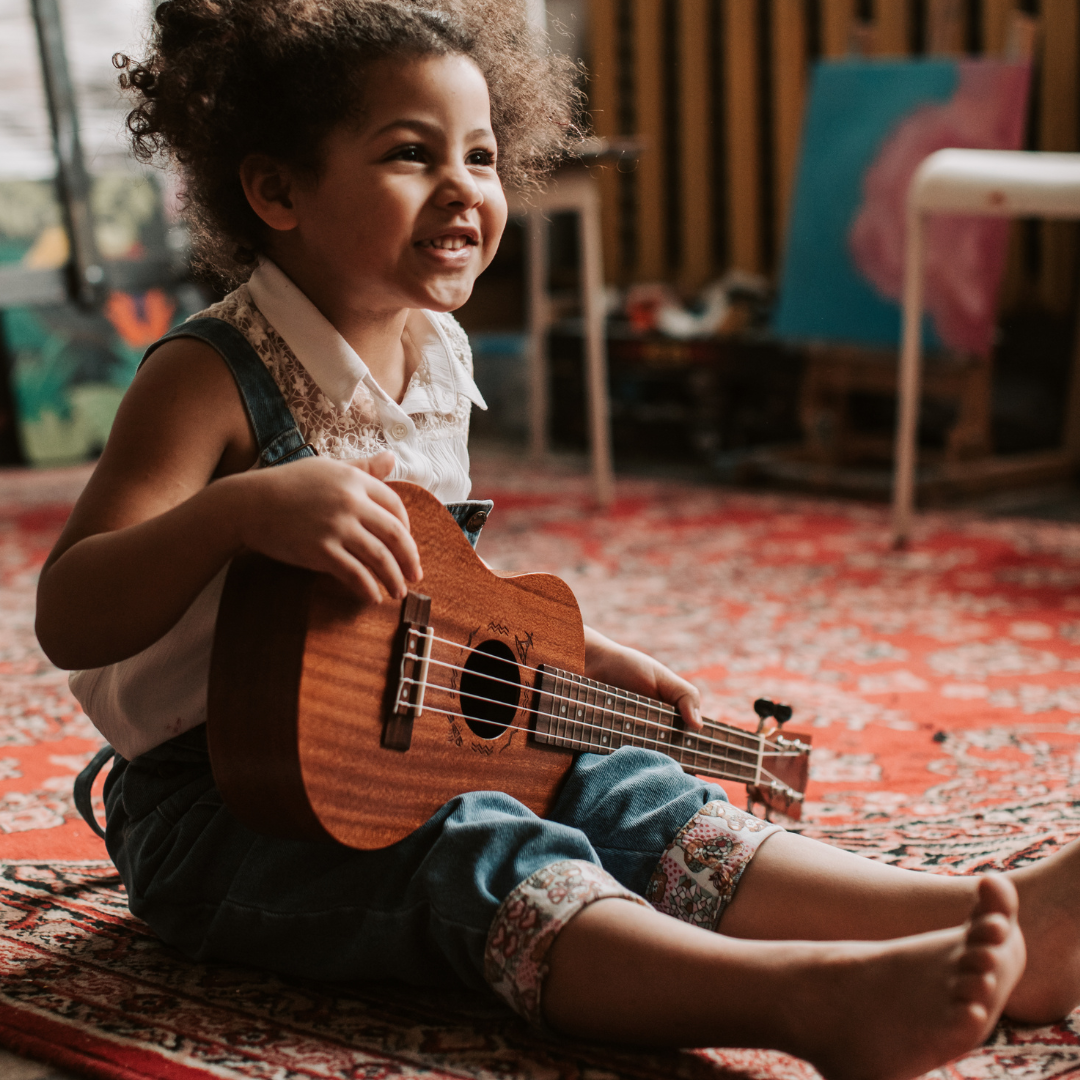 A little girl siting on the floor holding a guitar.