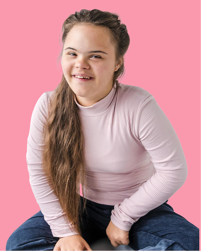 A young woman with long brown hair and Down syndrome smiles while sitting against a solid pink background. She is wearing a light pink ribbed long-sleeve top and blue jeans, embracing the joy promoted by music therapy Gold Coast sessions.