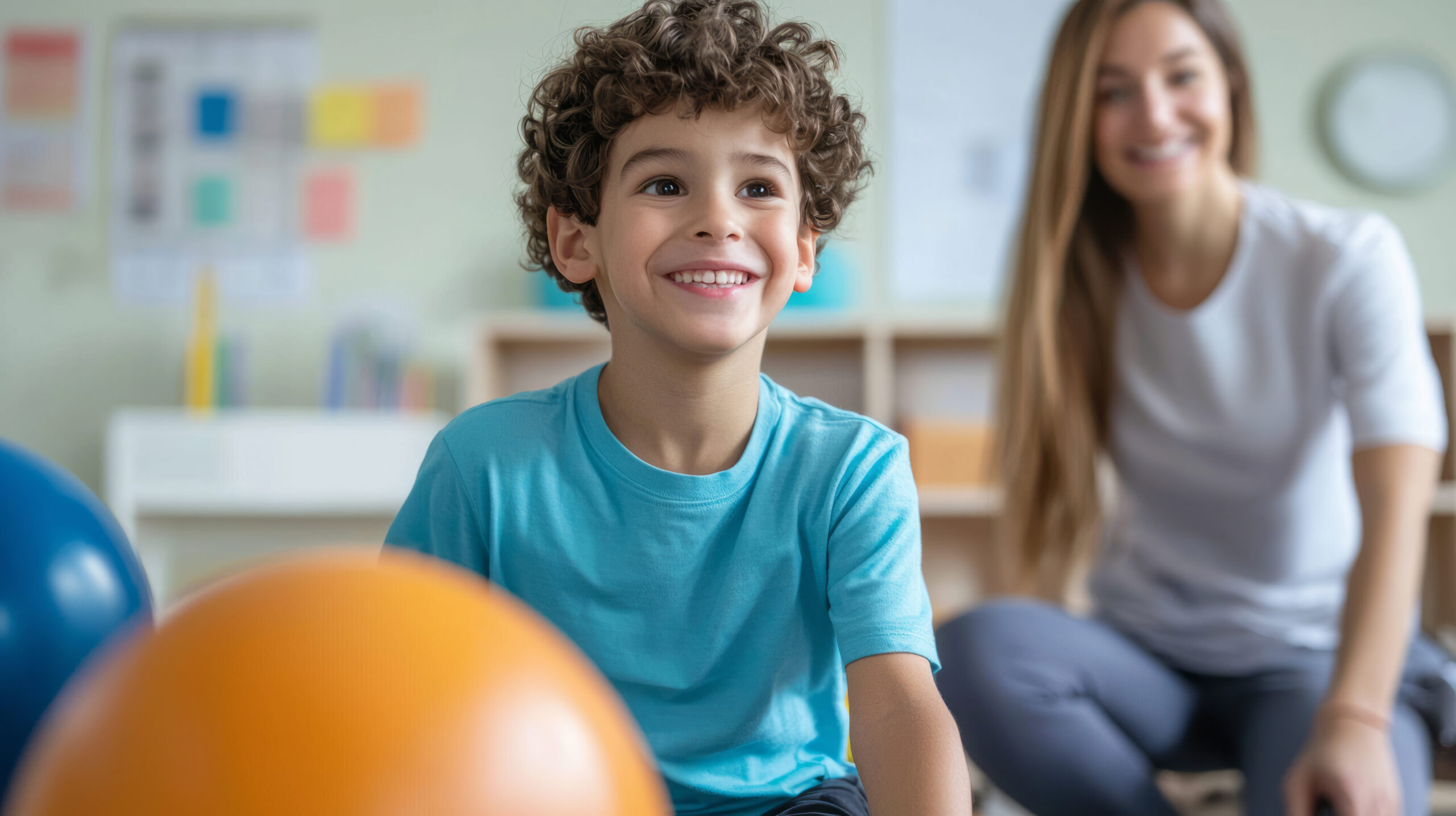 Smiling young boy in a blue shirt sits indoors with colourful balls in the foreground. A woman, likely a feeding therapist or carer, is seated and smiling in the blurred background.