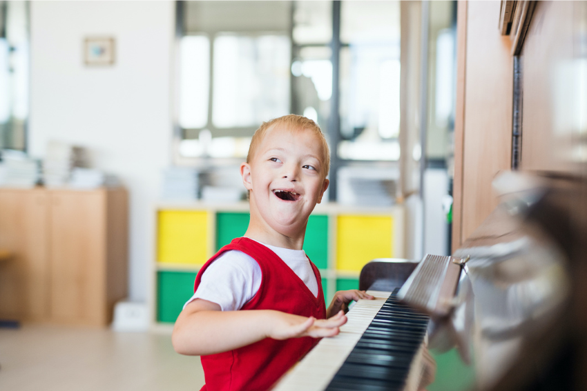 A young boy with Down’s syndrome smiles joyfully whilst playing the piano during a music therapy NDIS session in a bright room with colourful storage shelves in the background.