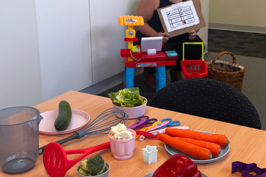 A table with vegetables, scissors, a whisk, and kitchen tools is in the foreground. In the background, an adult sits near a toy kitchen set, holding a visual communication board.