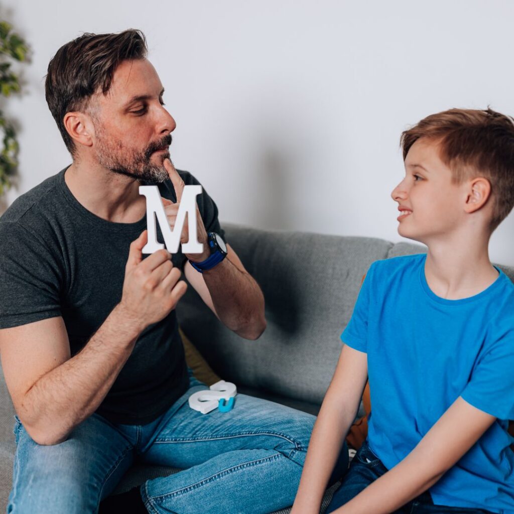 A man holding a white letter M to his lips sits on a sofa next to a smiling boy in a blue shirt, both looking at each other in a playful, relaxed setting.