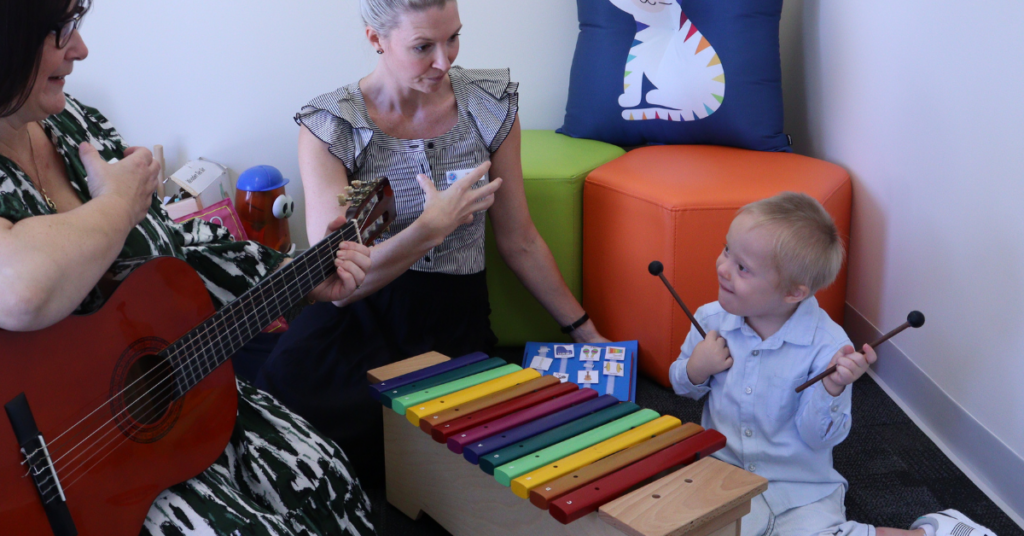 A music therapy with a guitar, a speech pathologist, both signing "more" to a young boy sitting on the floor with a xylophone.