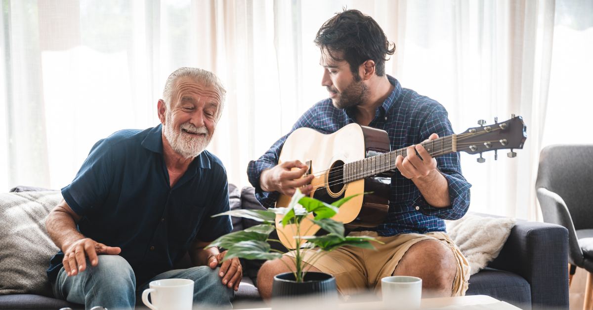 older man smiling and moving to music, sitting on a lounge with a male music therapist playing guitar.